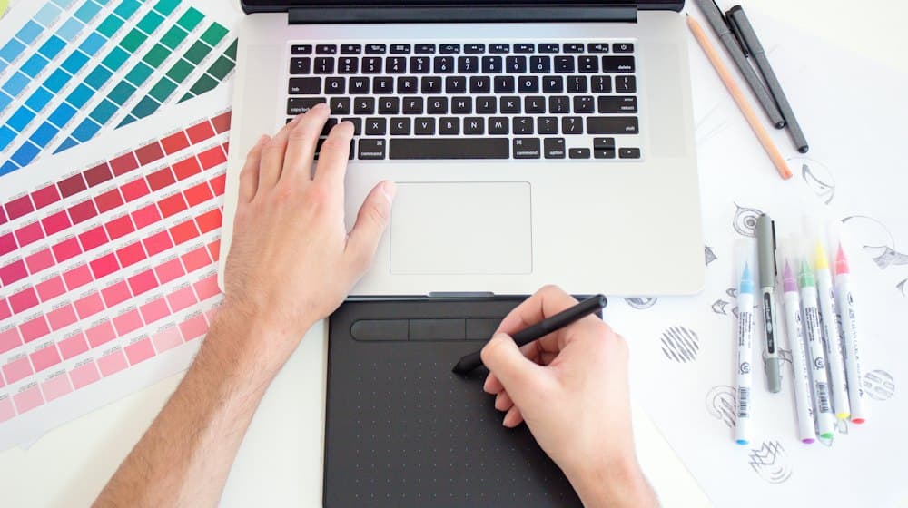 Un diseñador trabajando en el portátil junto a muestrarios de color y una tableta gráfica.