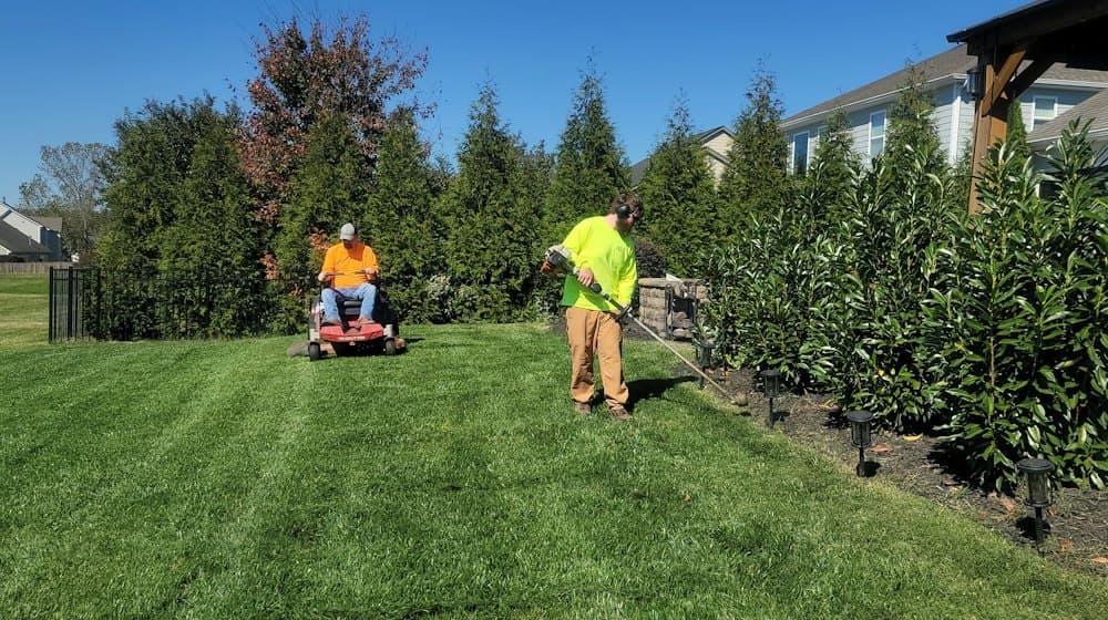 Two landscapers finishing a suburban back garden, one on a ride-on mower and one trimming the border.